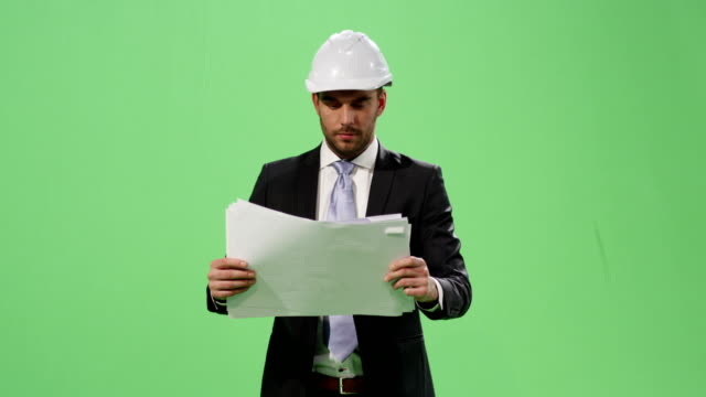 Empresario-en-un-casco-y-un-traje-es-caminar-y-buscar-en-documentos-en-papel-en-una-maqueta-pantalla-verde-de-fondo.