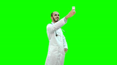 Sonriente-médico-de-bata-blanca-con-estetoscopio-tomando-selfie-en-su-teléfono-en-una-pantalla-verde-Chroma-Key
