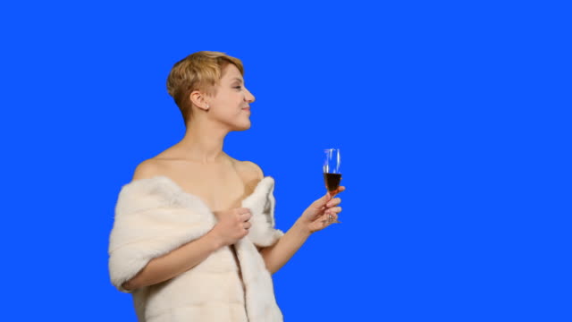 Charmante-Blondine-in-Pelz-Umhang-mit-roten-Wein-Posen-für-die-Kamera-auf-blauem-Hintergrund
