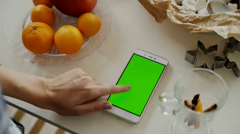 Closeup-of-woman's-hand-browsing-smartphone-with-green-screen-on-kitchen-table-at-home