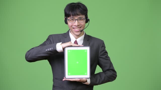 Junge-asiatische-Geschäftsmann-zeigt-digital-Tablette-zur-Kamera-während-seiner-Tätigkeit-als-Call-Center-Vertreter