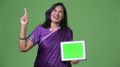 Mature-happy-beautiful-Indian-woman-thinking-while-showing-digital-tablet-and-pointing-finger-up