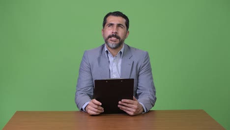 Handsome-Persian-bearded-businessman-thinking-while-holding-clipboard