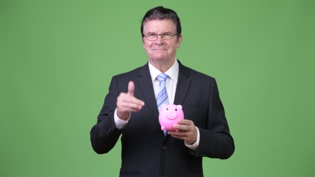 Senior-handsome-businessman-holding-piggy-bank-and-giving-thumbs-up