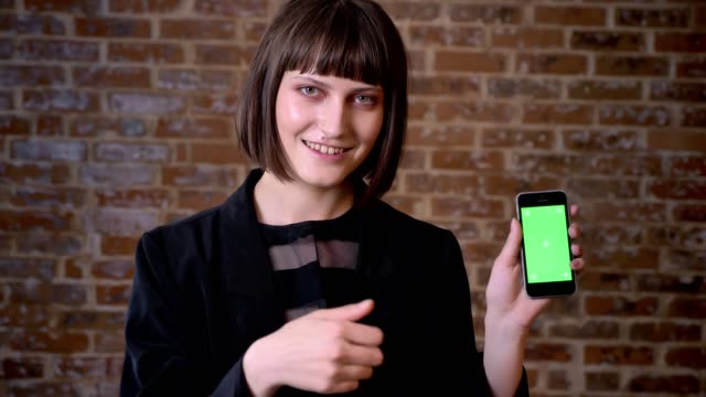 Young-happy-woman-holding-phone-with-chromakey-and-showing-thumbs-up,-brick-wall-background