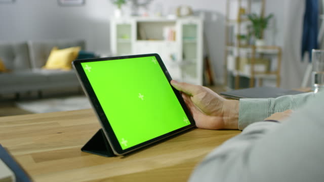 Over-the-Shoulder-Shot-of-a-Man-Holding-and-Watching-Green-Mock-up-Screen-Digital-Tablet-Computer-While-Sitting-at-the-Desk.-In-the-Background-Cozy-Living-Room.
