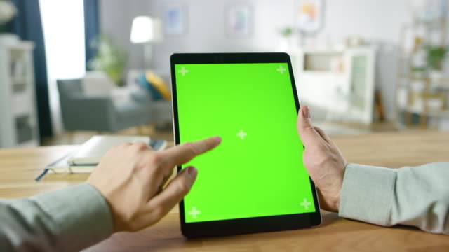 Close-up-of-Man-Using-Hand-Gestures-on-Green-Mock-up-Screen-Digital-Tablet-Computer-in-Portrait-Mode-while-Sitting-at-His-Desk.-In-the-Background-Cozy-Living-Room.