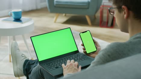 Young-Man-at-Home-Works-on-a-Laptop-Computer-with-Green-Mock-up-Screen,-while-Holding-Smartphone-with-Chroma-Key-Display.-He's-Sitting-On-a-Couch-in-His-Cozy-Living-Room.-Over-the-Shoulder-Camera-Shot