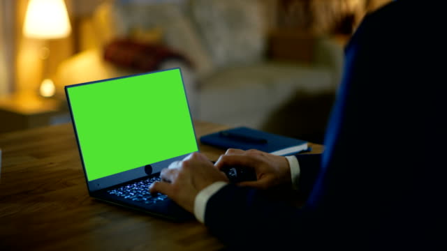 En-Home-Man-se-sienta-en-su-escritorio-y-tipos-en-un-ordenador-portátil-con-pantalla-verde-en-él.-Su-apartamento-está-hecho-en-colores-amarillos-y-es-cálido.