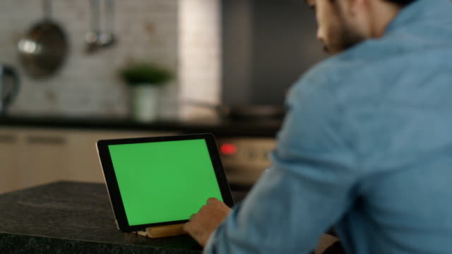 En-la-cocina.-Hombre-joven-sorbos-de-una-taza-durante-el-uso-de-Tablet-PC-con-pantalla-verde.