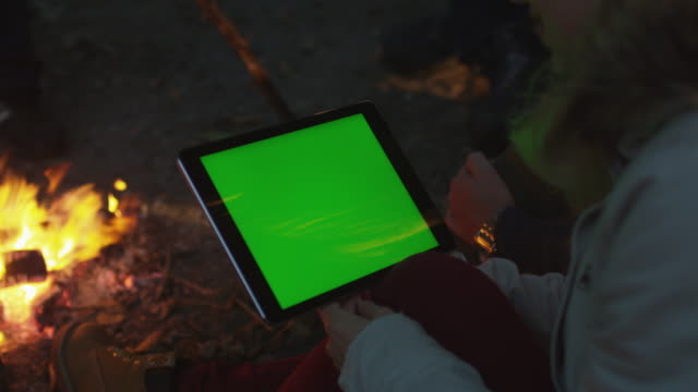 Mädchen-hält-einen-Tablet-im-Querformat-mit-Greenscreen-Mock-up-neben-einem-Lagerfeuer-am-Abend.