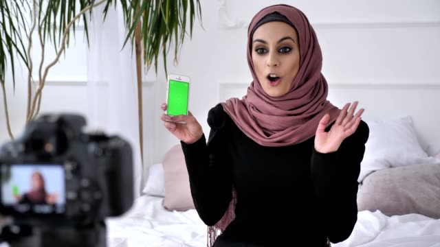 Young-beautiful-Indian-girl-in-hijab-recommends-an-application-on-a-smartphone,-smiling,-talking-at-the-camera,-chroma-key,-green-screen-concept.-50-fps