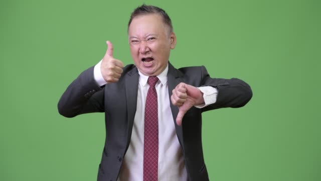 Mature-confused-Japanese-businessman-choosing-between-thumbs-up-or-thumbs-down