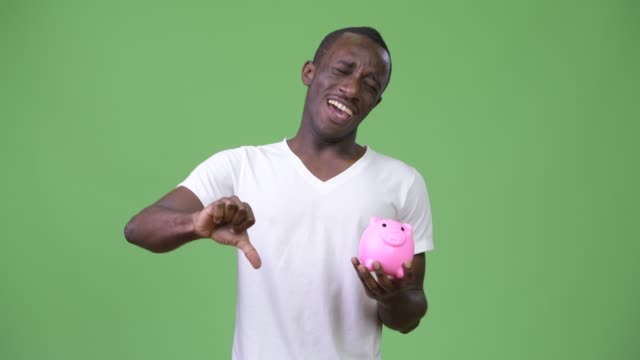 Young-African-man-holding-piggy-bank-and-giving-thumbs-down