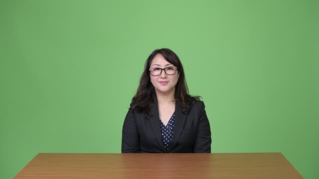 Reife-gerne-schöne-asiatische-geschäftsfrau-lächelnd-hinter-Schreibtisch-sitzend