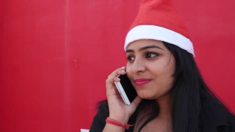 Handheld-shot-of-Indian-woman-with-Santa's-hat-talking-on-mobile-cell-phone,-cheerful-and-excited,-with-red-background