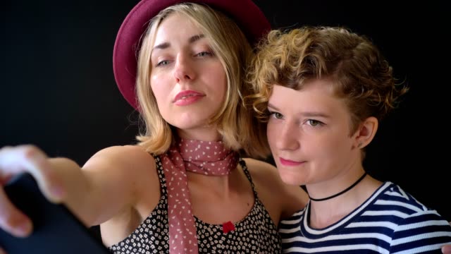 Zwei-charmante-stilvolle-junge-Frauen-mit-kurzen-Haaren-umarmen-und-nehmen-Selfie,-auf-schwarzem-Hintergrund-isoliert