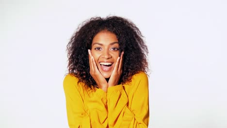 Beautiful-african-american-woman-with-afro-hair-in-yellow-wear-smiling,-pleasantly-surprised-to-camera-over-white-wall-background.-Cute-mixed-race-girl's-portrait-with-amazement