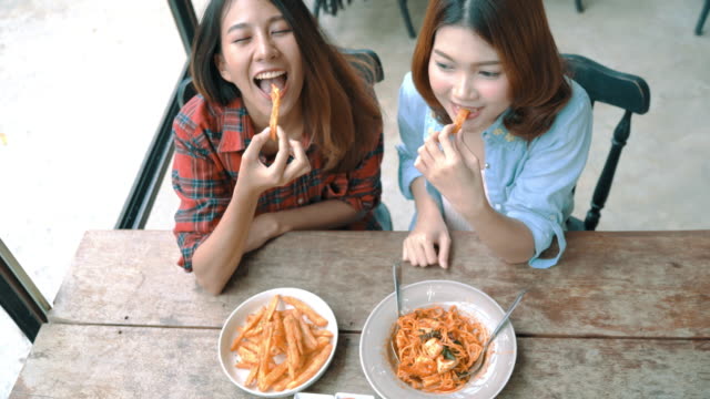 Beautiful-happy-Asian-women-lesbian-lgbt-couple-sitting-each-side-eating-a-plate-of-Italian-seafood-spaghetti-and-french-fries-at-restaurant-or-cafe-while-smiling-and-looking-at-food.