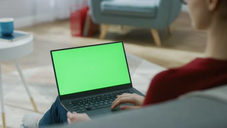 Junge-Frau-zu-Hause-arbeitet-an-einem-Laptop-Computer-mit-grünen-Mock-up-Bildschirm.-Sie-sitzt-auf-einer-Couch-in-seinem-gemütlichen-Wohnzimmer.-Über-die-Schulter-Kamera-Aufnahme