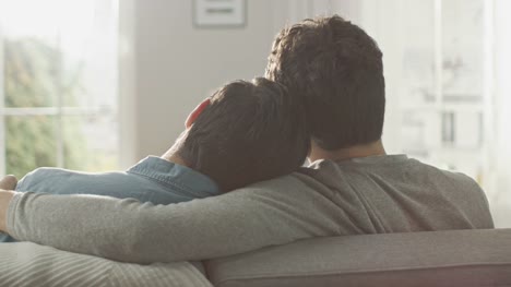 Back-Shot-of-Cute-Male-Queer-Couple-Hug-While-Sitting-on-a-Sofa-and-Putting-Their-Heads-Together.-They-are-Casually-Dressed-in-Jeans-and-Sweater.-Room-has-Modern-Interior-and-It's-Bright-and-Sunny.