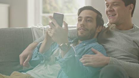 Cute-Male-Gay-Couple-Spend-Time-at-Home.-They-are-Lying-Down-on-a-Sofa-and-Use-a-Smartphone.-They-Browse-Online.-Partner's-Hand-is-Around-His-Lover.-They-Smile-and-Laugh.-Room-Has-Modern-Interior.