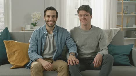 Cute-Attractive-Male-Gay-Couple-Sit-Together-on-a-Sofa-at-Home.-Boyfriend-Puts-His-Hand-on-Fiance's.-They-are-Happy-and-Smiling.-They-are-Casually-Dressed-and-Their-Room-Has-Modern-Interior.