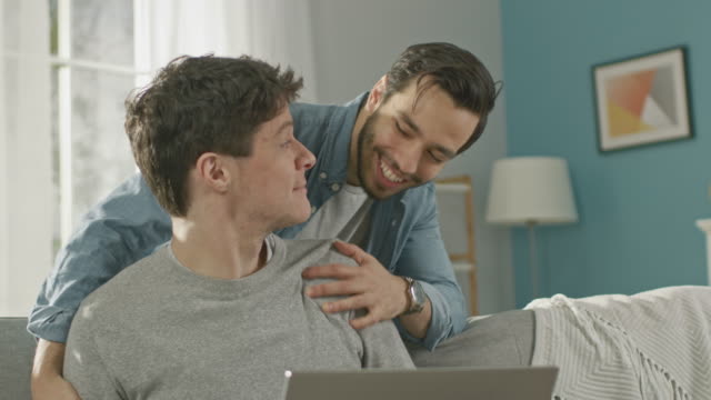 Sweet-Male-Gay-Couple-Spend-Time-at-Home.-Young-Man-Works-on-a-Laptop,-His-Partner-Comes-From-Behind-and-Gently-Embraces-Him.-They-Laugh-and-Touch-Hands.-Room-Has-Modern-Interior.