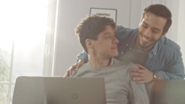 Dulce-hombre-Gay-pareja-pasar-tiempo-en-casa.-Hombre-joven-trabaja-en-un-ordenador-portátil,-su-compañero-viene-desde-atrás-y-suavemente-le-abraza.-Se-ríen-y-tocar-manos.-Habitación-tiene-un-Interior-moderno.