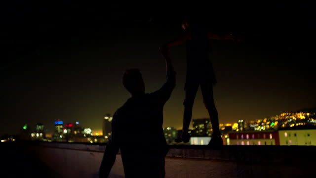Man-is-holding-hand-with-girlfriend-who-is-balancing-on-rooftop-edge
