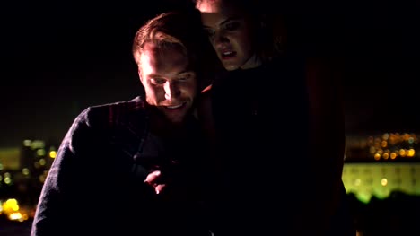 Young-couple-looking-at-smart-phone-on-rooftop-at-night