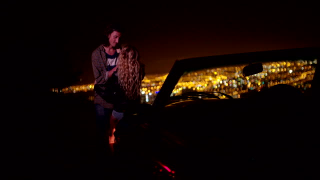 Young-couple-hugging-and-looking-at-night-city-lightsnext-to-a-convertible