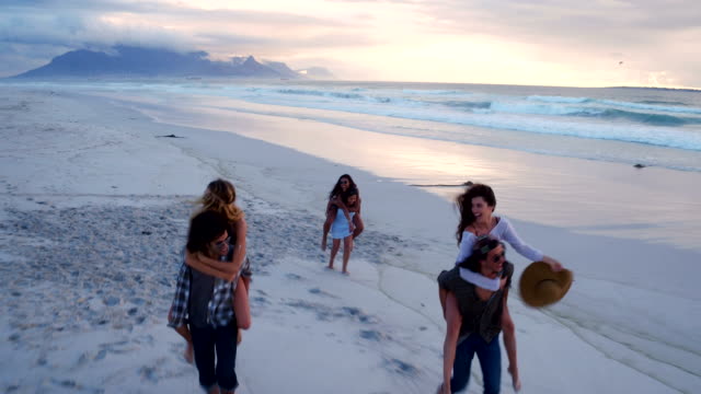 Hipster-friends-having-funny-piggyback-rides-on-beach-at-sunset
