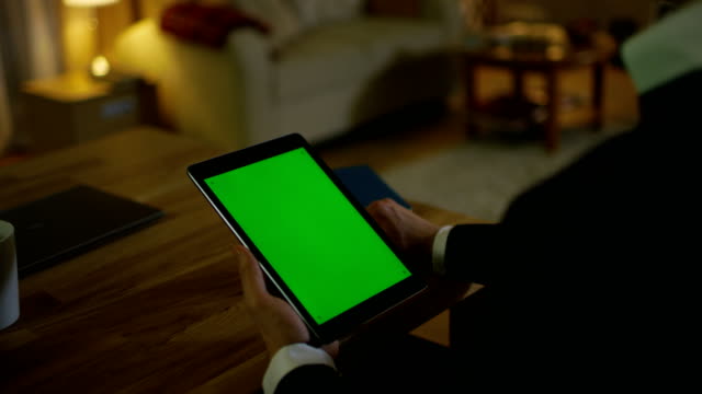 At-Home-Man-Sits-at-His-Desk-and-Holds-Tablet-Computer-with-Green-Screen-on-it.-His-Apartment-is-Done-in-Yellow-colours-and-is-Warm.