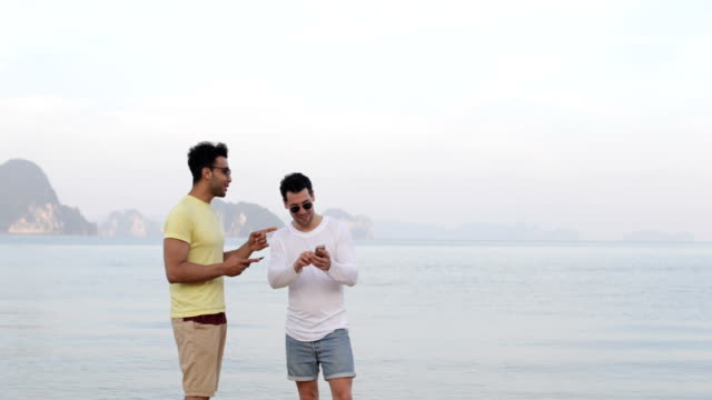 Dos-hombres-en-la-playa-usando-celular-teléfonos-hablando-de-Pareja-Gay-en-línea-de-redes-inteligentes