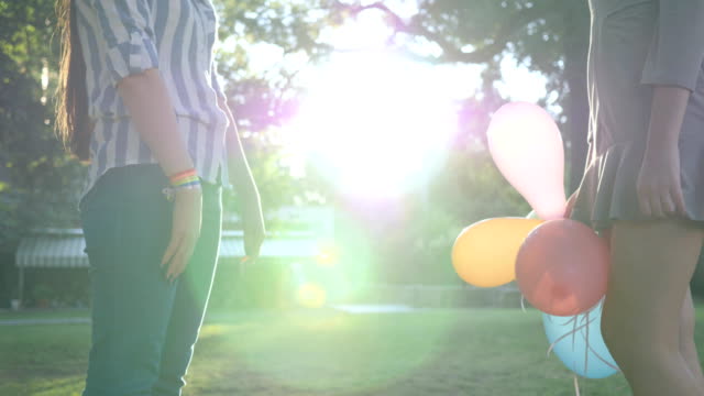 LGBT-girlfriends-stand-together-holding-hands-with-inflatable-balloons-and-bracelet-in-backlight
