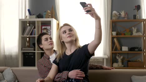 Zwei-junge,-schöne-Mädchen-sitzen-auf-der-Couch-und-machen-Selfies,-Lesben,-LGBT,-blond,-brünett-60fps