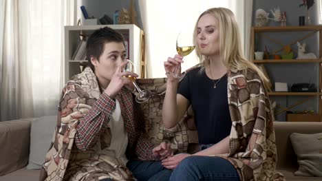 Two-young,-beautiful-girls-are-sitting-on-the-couch-wrapping-with-warm-plaid-blanket,-clink-glasses-and-drink-60-fps