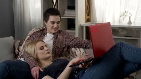 Young-blond-woman-lies-on-the-lap-of-a-brunette-and-using-red-laptop,-cosiness,-cuddles,-plaid-60-fps