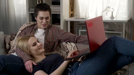 Young-blond-woman-lies-on-the-lap-of-a-brunette-and-using-red-laptop,-cosiness,-cuddles,-plaid,-laughing-60-fps