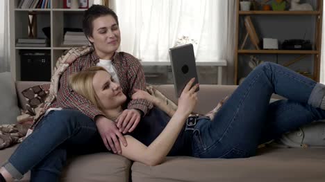Lesbisches-Paar-ruht-sich-auf-der-Couch,-mit-Tablet-PC,-Mädchen-mit-kurzen-Haaren-sanft-streichelt-ihr-Partner-Gesicht-sprechen,-Familienidylle,-LGBT-60-fps