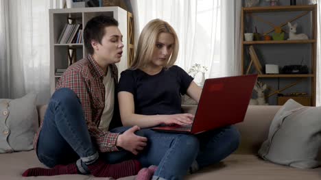 Dos-jóvenes-lesbianas-niñas-están-sentados-en-el-sofá,-usando-una-computadora,-internet,-compras-online,-la-rubia-maliciosa-de-desplazamiento-cierra-la-laptop,-pelean-60-fps