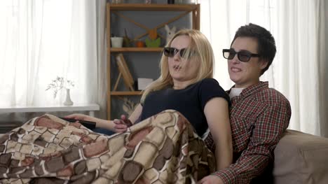 Dos-jóvenes-lesbianas-están-sentados-en-el-sofá,-cubierto-de-una-manta,-viendo-la-película-en-3d,-usando-el-control-remoto,-sorprendido,-sorprendido-60-fps