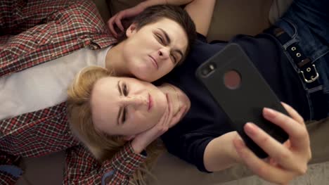 Two-young-girls-lie-on-the-couch,-do-selfie-on-a-smartphone,-grimace,-make-funny-faces,-top-shot-60-fps