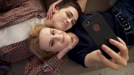 Two-young-girls-lie-on-the-couch,-do-selfie-on-a-smartphone,-smiling,-lovers,-lgbt,-young-couple,-top-shot-60-fps