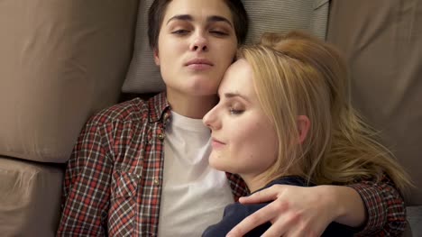 Two-young-lesbian-girls-lie-on-the-couch,-hug,-cuddle,-sleep,-girl-with-short-hair-looks-at-the-camera,-lgbt-family-concept,-top-shot-60-fps