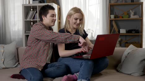 Dos-jóvenes-lesbianas-están-sentados-en-el-sofá,-usando-una-computadora,-internet,-compras-en-línea,-sonriendo,-hablando-de-desplazamiento