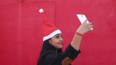 Indian-woman-with-Santa's-hat-taking-selfies-photo-with--mobile-cell-phone,-cheerful-and-excited,-with-red-background