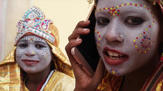 Handheld-close-up-of-young-girls-dressed-as-Hindu-God-talking-communication-technology-on-a-smart-mobile-phone-and-look-at-the-camera-happy-joy-content