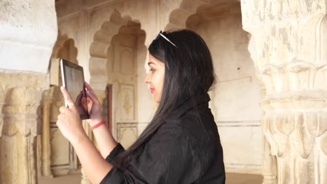 Beautiful-woman-with-black-eyes-taking-photos-and-working-on-a-touch-screen-tablet-pad-handheld-business-network-online-wireless-connection-at-an-old-building-complex-ancient-Indian-temple-column